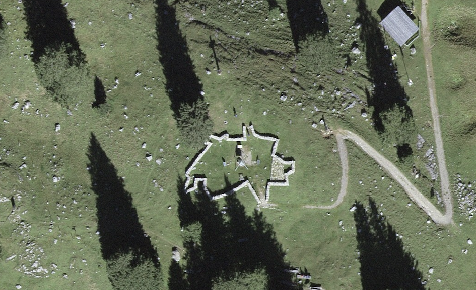 An aerial satellite view of a grassy alpine meadow. At the center is a large, low, jagged stone wall structure that precisely traces the outline shape of Switzerland's border. Inside the shape are some small triangular markers and a public fire pit/BBQ area. The structure is surrounded by high-altitude pine trees and a small, lone wooden building to the upper right.